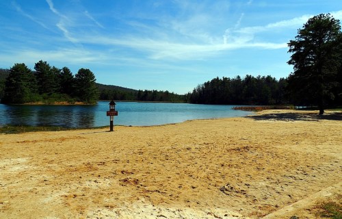 A sandy beach by a calm lake, surrounded by trees and blue skies with wispy clouds.