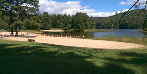 A sandy beach by a calm lake, surrounded by trees and a blue sky with clouds. Colorful flags line the beach area.
