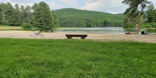 A sandy beach by a calm lake, surrounded by green trees and hills, with people relaxing and swimming.