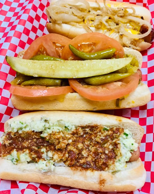 Three hot dogs on a red and white checkered tray: one with chili and toppings, one with pickles and tomatoes, and one with onions and cheese.
