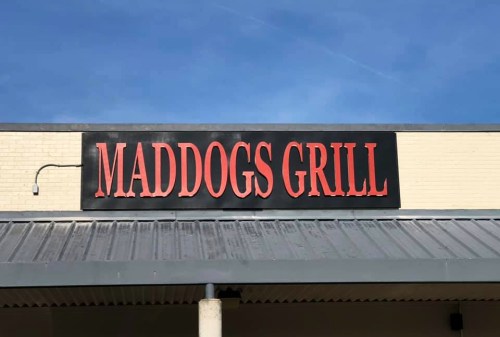 Sign for "Maddogs Grill" against a blue sky, featuring bold red letters on a light-colored background.