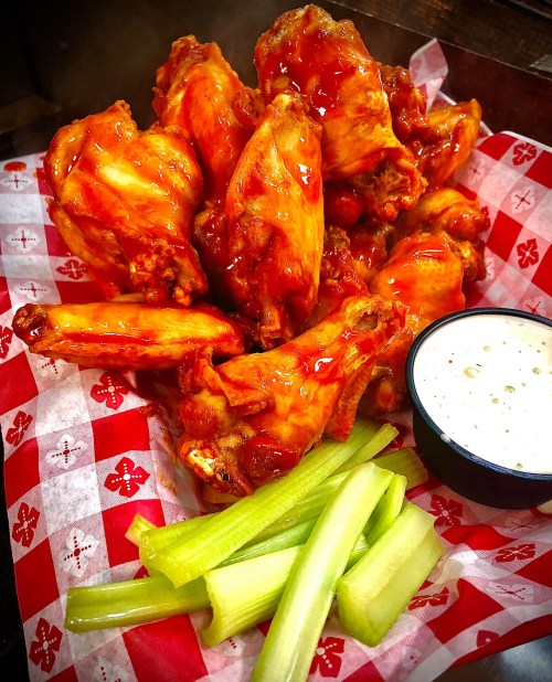 A pile of saucy chicken wings on a red and white checkered paper, served with celery sticks and a small bowl of ranch dressing.