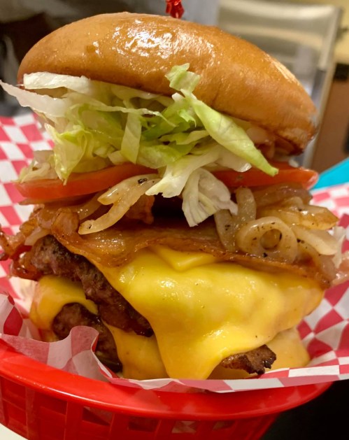 A towering cheeseburger with lettuce, tomato, grilled onions, and bacon, served in a red basket with checkered paper.