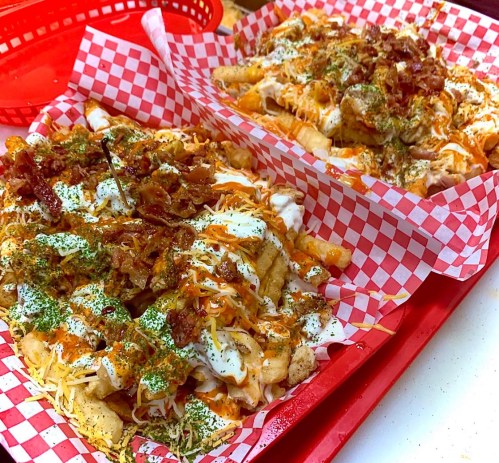 Two trays of loaded fries topped with chicken, cheese, bacon, and drizzled sauces, served in red checkered paper.
