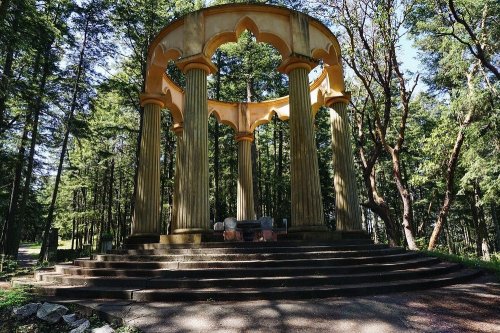 A classical-style gazebo with columns, surrounded by tall trees in a forest setting. Sunlight filters through the leaves.