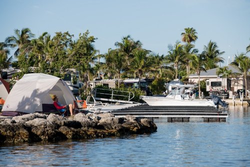 A serene waterfront scene with a tent, a person relaxing, and boats docked among palm trees.