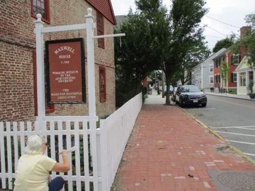 A woman kneels by a sign for Maxwell House, a historic museum, on a brick sidewalk lined with trees and buildings.