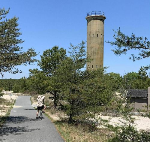 A cyclist rides on a path near a tall, cylindrical concrete tower surrounded by trees and sandy terrain.
