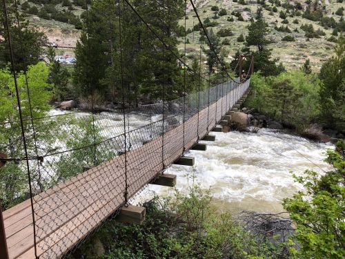 A suspension bridge spans a rushing river, surrounded by lush greenery and rocky terrain in a mountainous area.