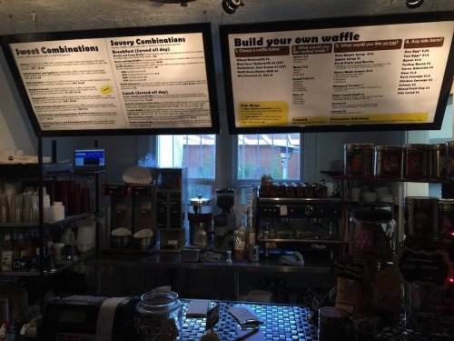 Menu boards displaying sweet and savory waffle combinations in a café setting, with a coffee machine in the background.