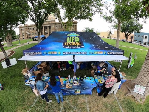 A colorful tent at the UFO Festival in Roswell, New Mexico, with people browsing merchandise under a tree.