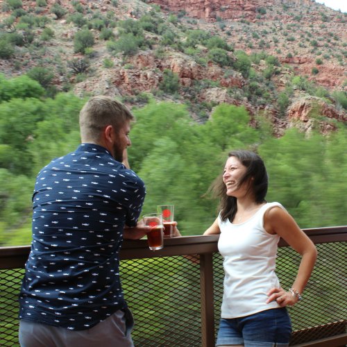 A man and woman enjoy drinks on a balcony with a scenic view of green trees and red rock formations.