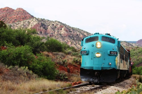 A vintage turquoise train travels along a scenic route through rugged mountains and greenery.