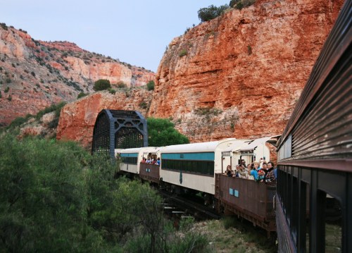 A vintage train travels along a scenic route, passing through rocky terrain and a metal bridge with passengers visible.