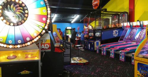 A vibrant arcade with colorful games, a spinning wheel, and people playing in a lively atmosphere.