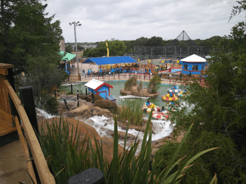 A colorful water park scene with pools, slides, and people enjoying various water activities amidst greenery.