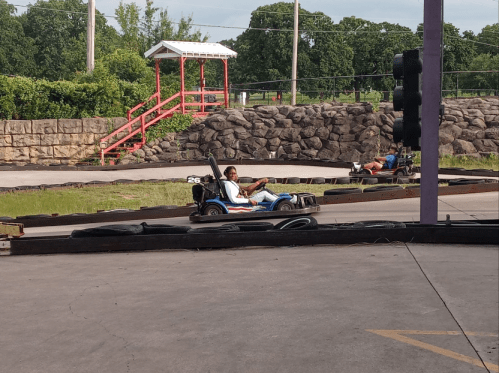 A go-kart track with two karts racing; one driver is focused while the other is in the background. Green trees in view.