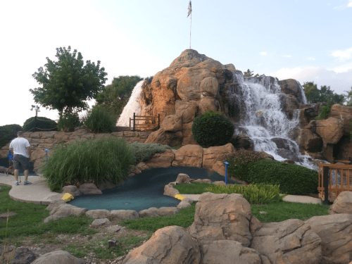 A scenic waterfall cascading over rocks, surrounded by greenery and a small pool, with a person walking nearby.