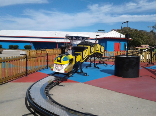 A child rides a colorful, cartoonish train on a small amusement park track under a blue sky.