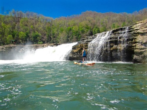 A person stands on a paddleboard in front of a waterfall, surrounded by lush green trees and a clear blue sky.