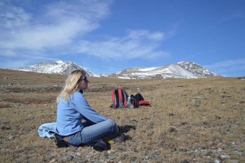 A woman sits on a grassy hillside, surrounded by mountains under a clear blue sky, with backpacks nearby.