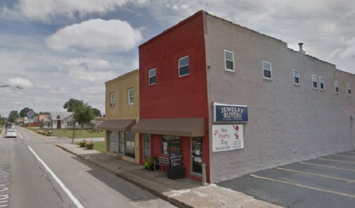 A small jewelry store with a red and gray exterior, located on a street with grassy areas and parked cars nearby.