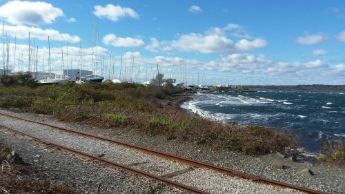 A coastal scene with a rocky shore, boats in the distance, and a railway track lined with shrubs under a blue sky.
