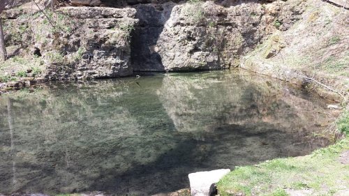 A serene pond surrounded by rocky cliffs and greenery, reflecting the landscape above.