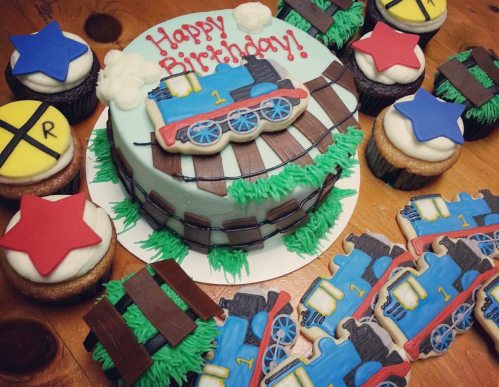 A colorful birthday cake with a train design, surrounded by themed cupcakes and cookies featuring train and star decorations.