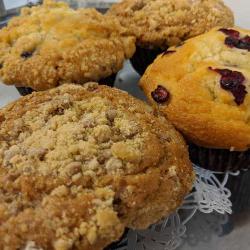 Four freshly baked muffins with crumbly tops, featuring flavors like blueberry and cranberry, arranged on a decorative tray.