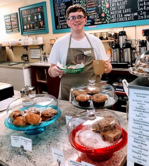 A smiling barista holds a plate of cookies and a cup in a cozy café with baked goods displayed in glass domes.