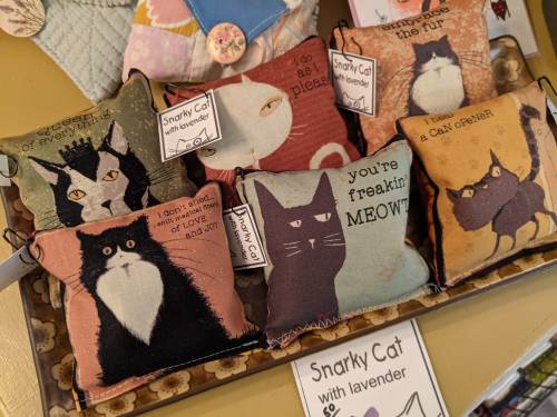 A display of colorful cat-themed pillows with humorous phrases, featuring various cat illustrations and lavender scent.