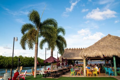A vibrant outdoor restaurant by the water, featuring colorful chairs, palm trees, and a thatched roof.
