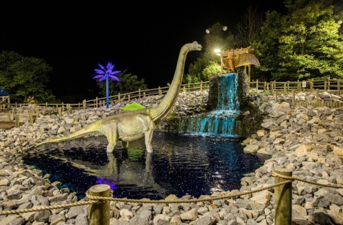 A dinosaur statue near a waterfall and pond, surrounded by rocks and illuminated by colorful lights at night.
