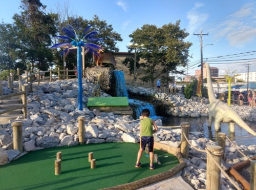 A child plays mini-golf near a waterfall and palm trees, with a dinosaur statue in the background.