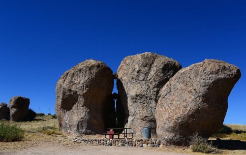 Two large boulders stand side by side, creating a natural arch, with a picnic table nearby under a clear blue sky.