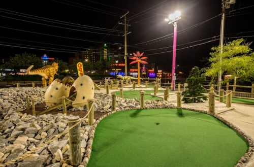 A nighttime view of a mini-golf course featuring dinosaur decorations, palm trees, and colorful lights in the background.