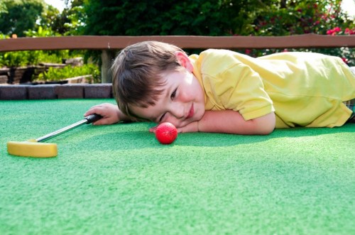 A young boy lies on a mini-golf course, resting his head on his arms next to a golf ball, with a putter nearby.