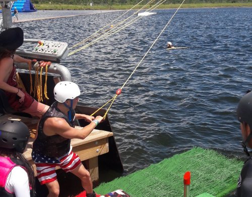 A person in a helmet prepares to wakeboard while others watch from a dock by a lake on a sunny day.