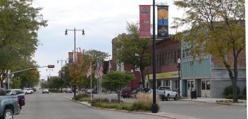 A quiet downtown street lined with trees, banners, and parked cars, featuring storefronts and American flags.