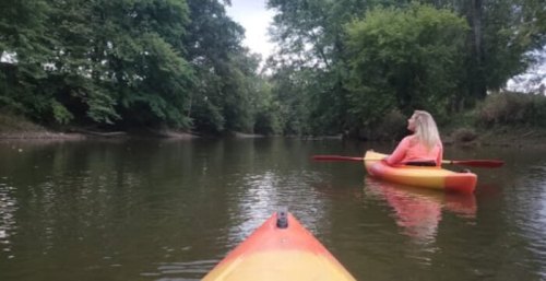 Two kayakers on a calm river, surrounded by lush greenery, with one person facing away in a bright orange kayak.
