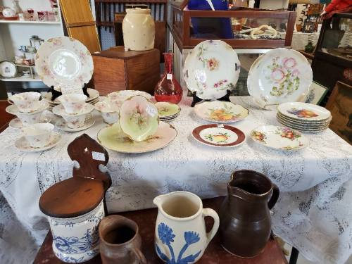A display of vintage china, including plates, cups, and pitchers, arranged on a lace tablecloth in an antique shop.
