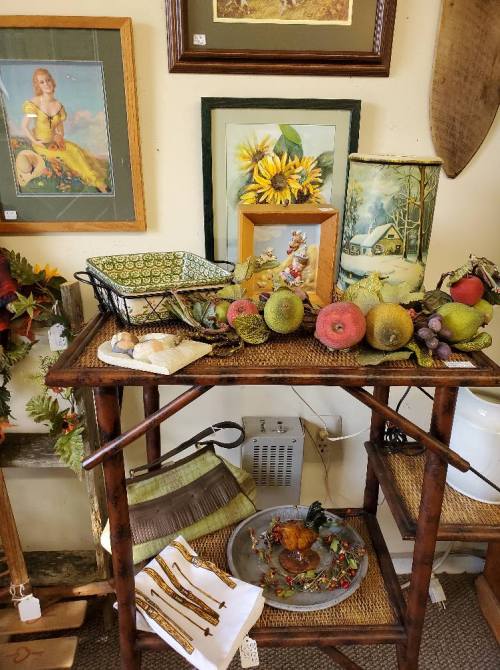A wooden table displays decorative items, including fruit, a basket, and framed art, in a cozy shop setting.