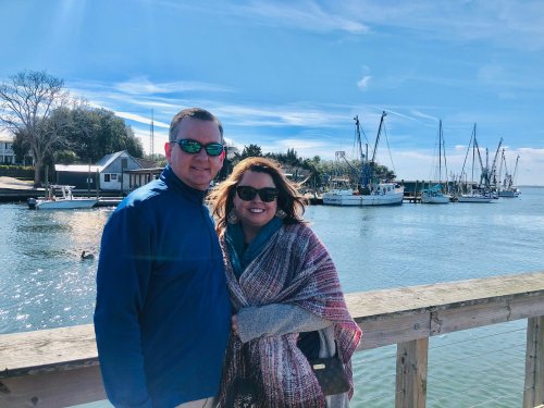 A couple stands on a pier by a harbor, smiling with boats and a clear blue sky in the background.
