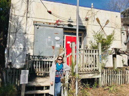 A woman stands in front of a weathered building with a red door and wooden steps, surrounded by greenery.