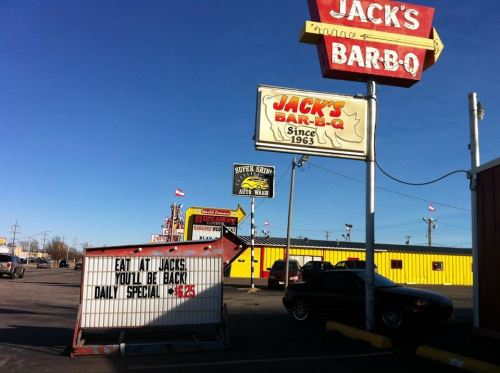 A roadside view of Jack's Bar-B-Q with a sign advertising daily specials and nearby businesses under a clear blue sky.