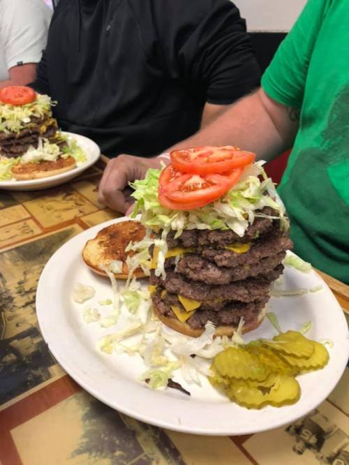 A towering burger stacked with multiple beef patties, cheese, lettuce, tomatoes, and pickles on a plate.