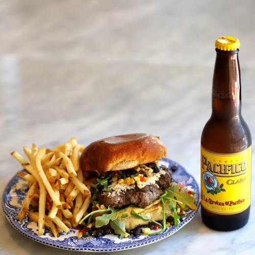 A juicy burger with toppings on a plate, accompanied by fries and a bottle of Pacifico beer.