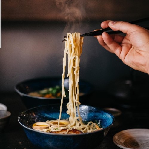 A hand holding chopsticks lifts steaming noodles from a blue bowl, with a second bowl of soup in the background.