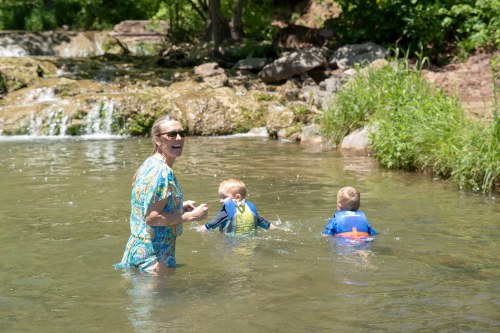 A woman smiles while wading in a stream with two young children, surrounded by greenery and rocks.